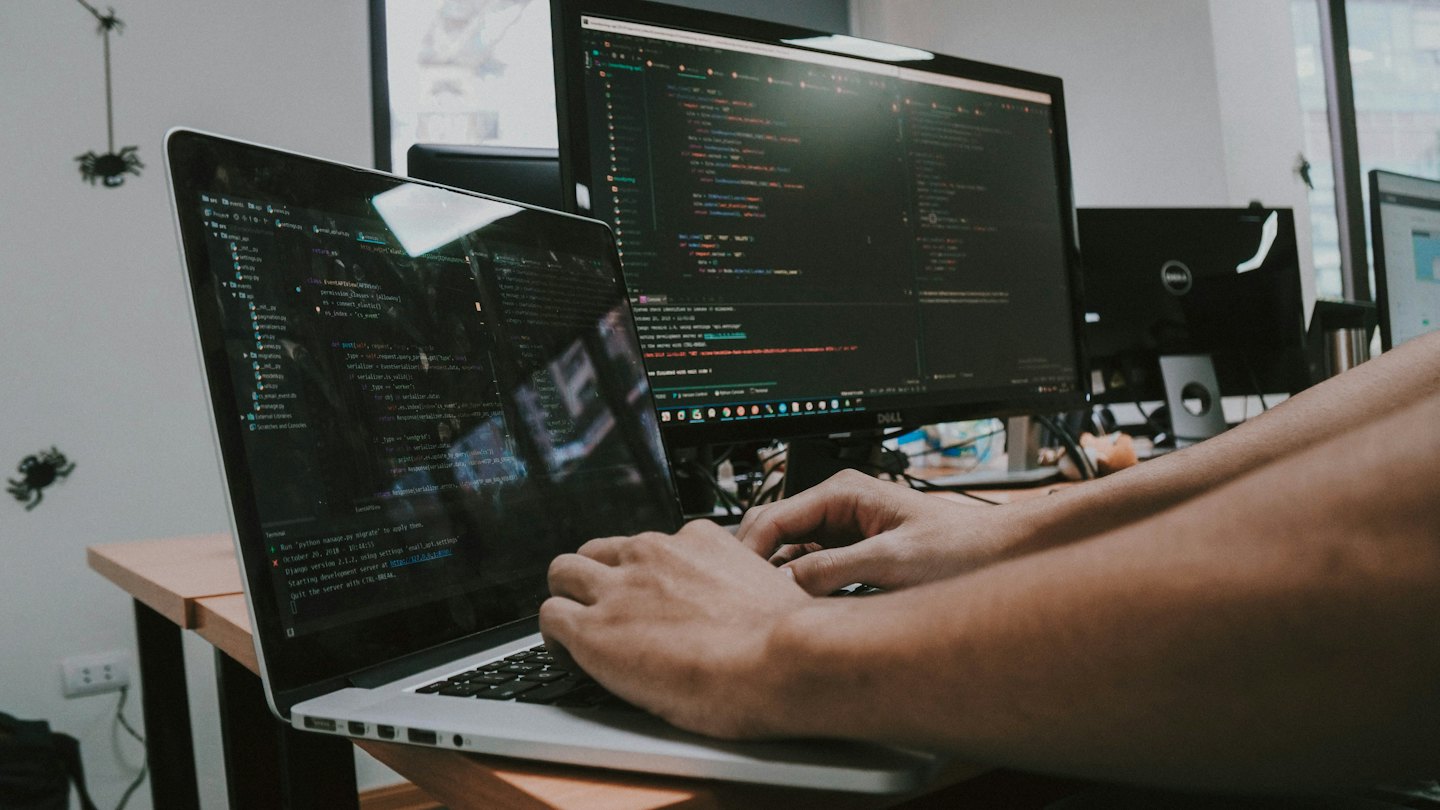 Manos escribiendo código en un portátil conectado a un monitor grande.