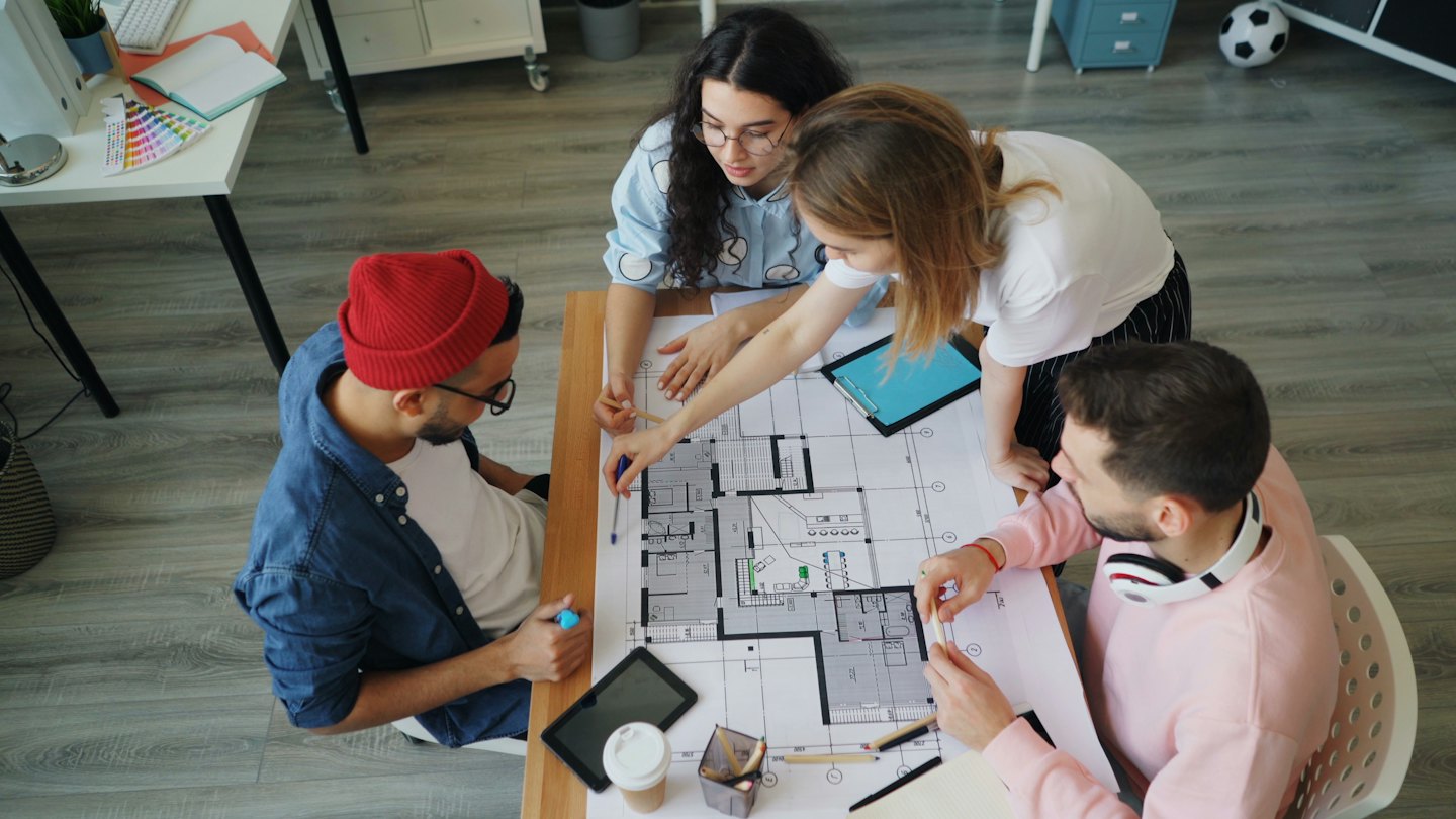 Cuatro diseñadores colaboran sobre un plano arquitectónico en una mesa.